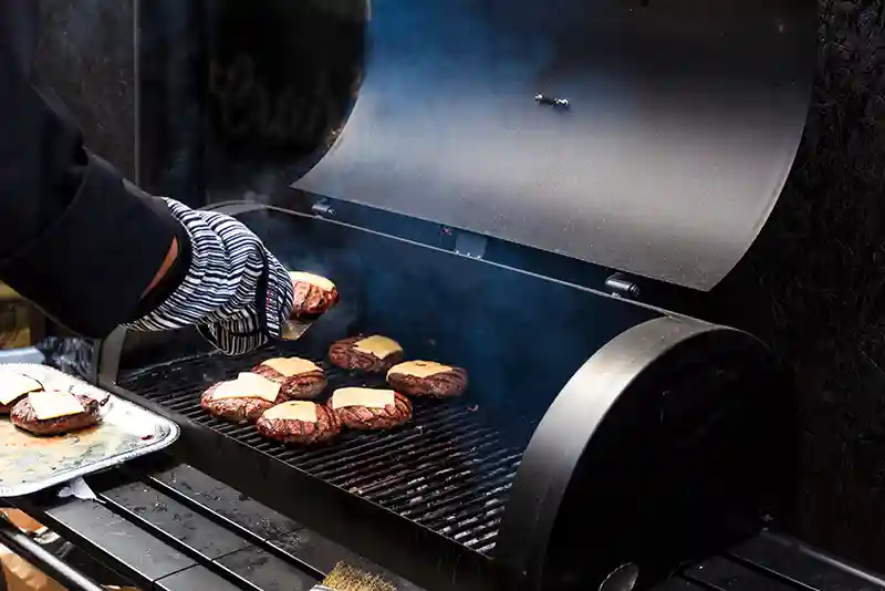 Auf einen schwarzen Holzkohlengrill werden Burgerpatties mit K&auml;se gelegt