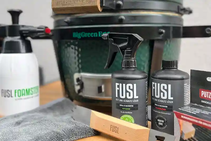 Verschiedene Reiniger und Reinigungszubeh&ouml;r stehen vor einem gr&uuml;nen Keramikgrill