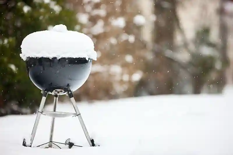 Ein schwarzer Kugelgrill mit einer Haube aus Schnee steht in einem verschneiten Garten